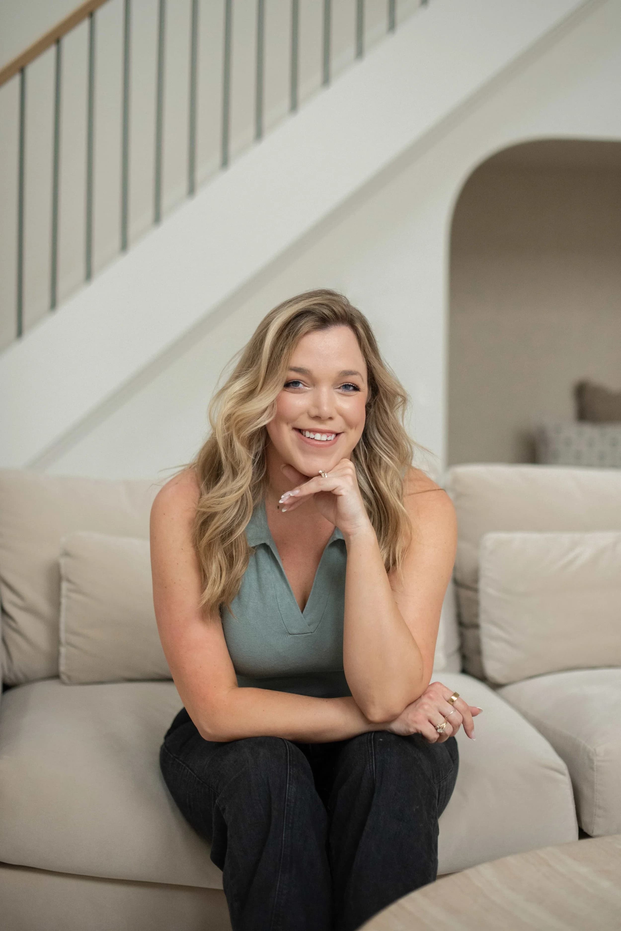 Kaitlin Kirby seated on a cream sofa beneath a curved staircase, smiling