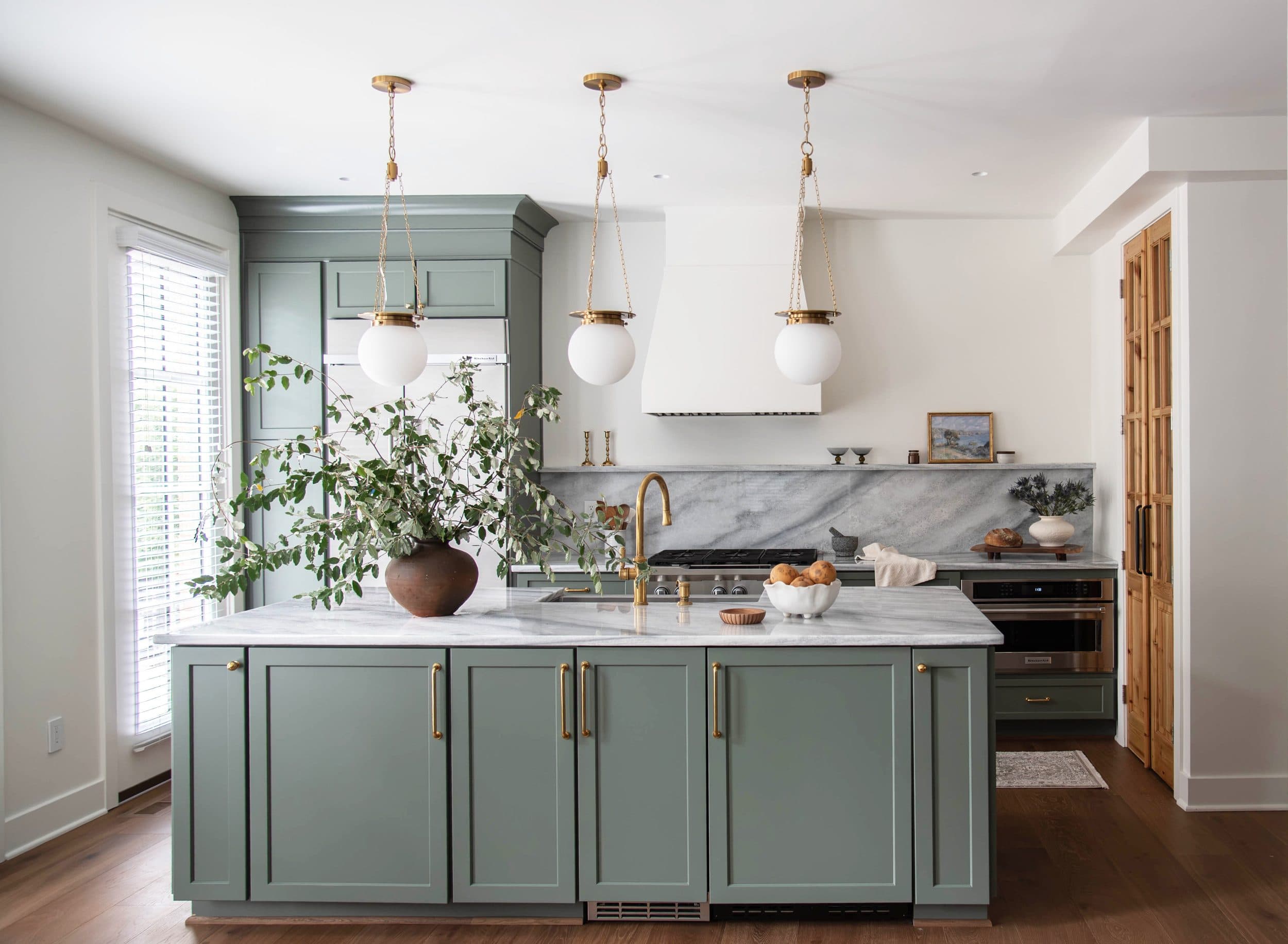 Sage green kitchen with a marble island, three brass and opal globe pendants, and a plaster range hood