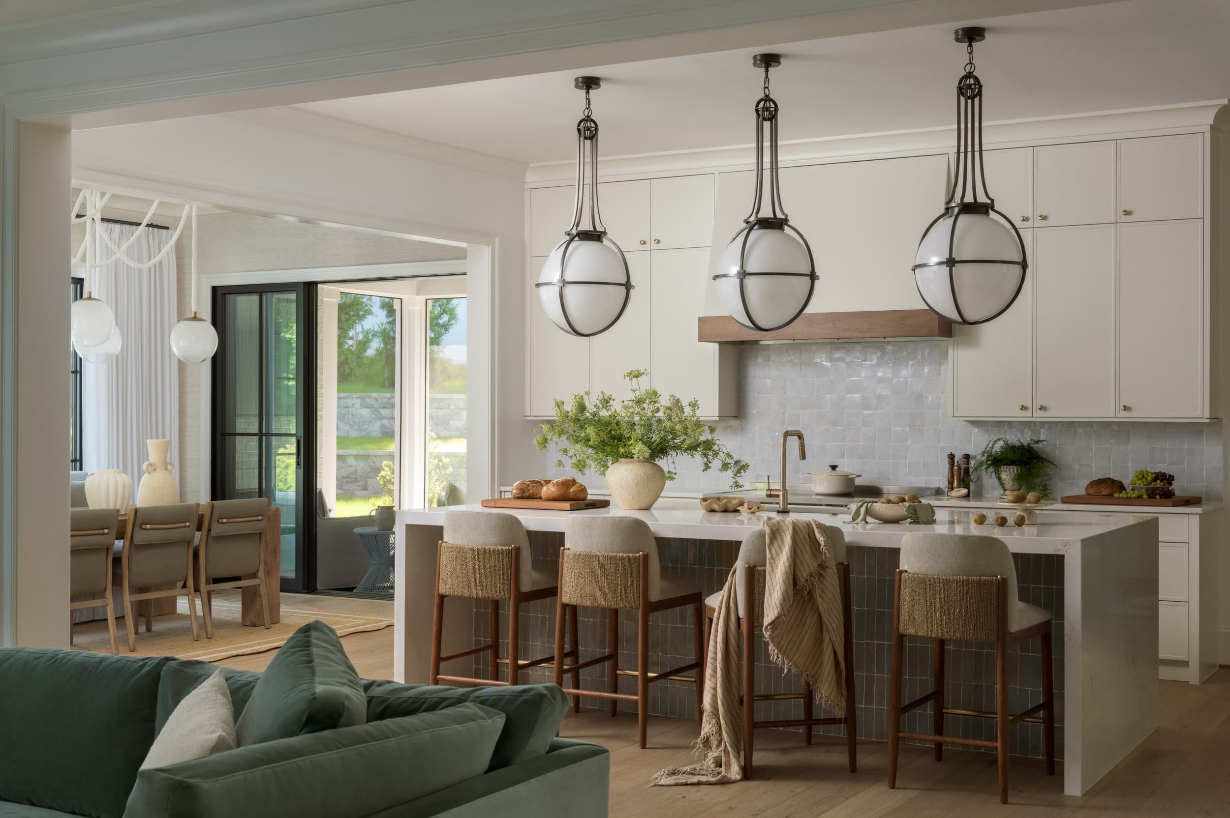 Open kitchen with globe pendants over a zellige-tiled island, woven counter stools, and a dining nook beyond